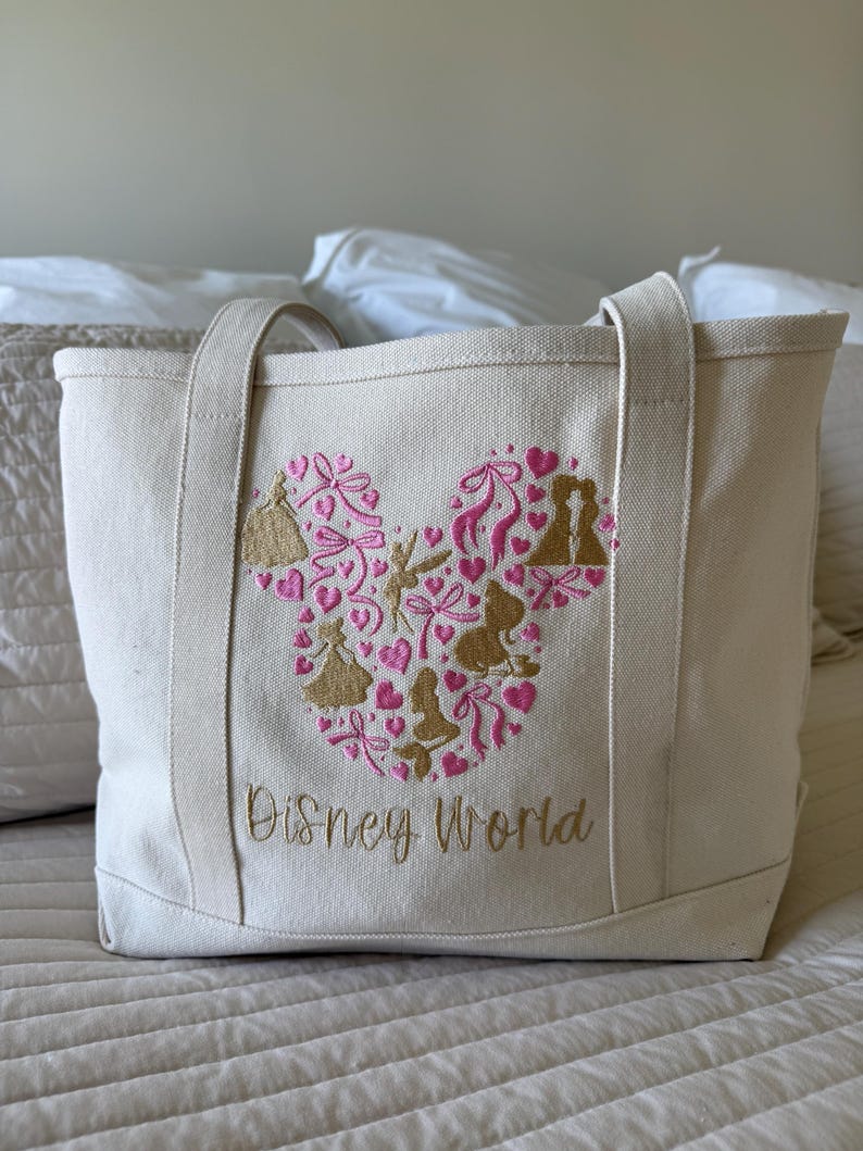 May include: Cream-colored canvas tote bag with embroidered design. The design features a Mickey Mouse head shape filled with pink hearts, bows, and gold silhouettes. The words "Disney World" are embroidered in gold below the design. The bag has two handles.