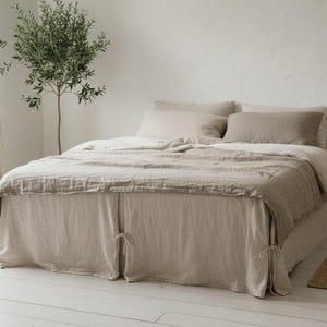 Może przedstawiać: Łóżko z pościelą lnianą w neutralnych odcieniach, w tym poszwą na kołdrę, poduszkami i spódnicą łóżka. Łóżko znajduje się w jasnym pokoju z małym drzewkiem i drewnianym stolikiem nocnym. Ogólna estetyka jest minimalistyczna i naturalna.