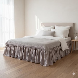 Może przedstawiać: Łóżko z jasnoszarą lnianą poszwą na kołdrę i pasującą spódnicą do łóżka. Łóżko jest pościelone białymi poduszkami i jasnoszarą poduszką lędźwiową. Wezgłowie jest w neutralnym beżowym kolorze. Łóżko stoi na drewnianej podłodze.