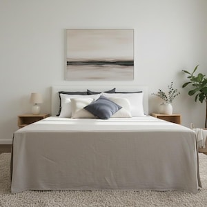 Może przedstawiać: Wnętrze sypialni z łóżkiem z pościelą w neutralnych odcieniach i poduszkami w kolorze białym i granatowym. Obraz pejzażowy wisi nad łóżkiem. Drewniane stoliki nocne z lampami po obu stronach. Roślina doniczkowa i wiklinowy kosz dopełniają wystroju.