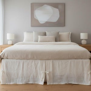 Może przedstawiać: Łóżko z beżową kołdrą i białą spódnicą łóżka z wiązanymi detalami. Łóżko jest zaopatrzone w kilka poduszek w odcieniach beżu i bieli. Dwie białe lampy stoją na drewnianych szafkach nocnych. Nad łóżkiem wisi duży abstrakcyjny obraz.