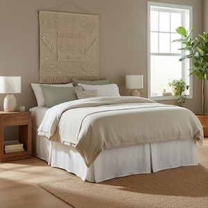 Może przedstawiać: Sypialnia z łóżkiem z beżową kołdrą i białą spódnicą łóżka. Tkany gobelin ścienny z geometrycznymi wzorami wisi nad łóżkiem. Dwie białe lampy stoją na drewnianych szafkach nocnych. Widoczne jest duże okno i roślina doniczkowa.