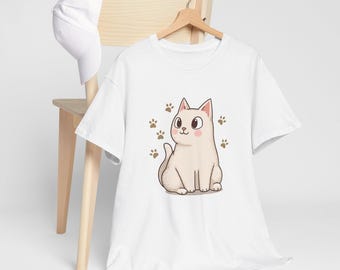 May include: White t-shirt featuring a cartoon cat illustration with brown paw prints. The cat is light beige with pink cheeks and a smiling expression. A white baseball cap rests on a wooden chair in the background.