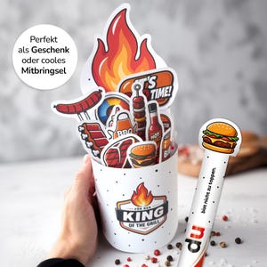 Könnte beinhalten: Ein weißer Becher mit Flamme und "KING OF THE GRILL"-Logo, gefüllt mit Aufklebern zum Thema Grillen, darunter Burger, Steak und Wurst. Ein Stab mit einem Burger und dem Text "bist nicht zu toppen" befindet sich an der Seite.