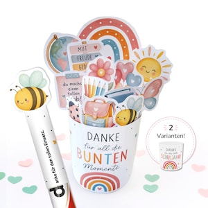 Könnte beinhalten: Ein weißer zylindrischer Behälter mit dem Text "DANKE für all die BUNTEN Momente" und einem Regenbogen-Motiv, gefüllt mit Schulausschnitten wie Globus, Stiften, Rucksack und Biene. Ein passender weißer Stift trägt die Aufschrift "anke für den tollen Einsatz."