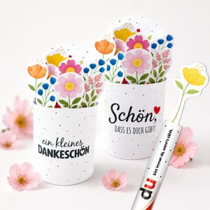 Könnte beinhalten: Zwei weiße zylindrische Behälter mit schwarzen Punkten, gefüllt mit bunten Papierblumen-Ausschnitten. Ein Behälter trägt die Aufschrift "ein kleines DANKESCHÖN", der andere "Schön, DASS ES DICH GIBT!". Ein separater weißer Stab zeigt eine gelbe Blume und Text.