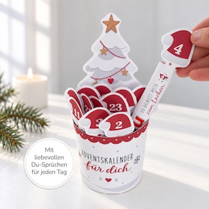 Könnte beinhalten: Ein Adventskalender aus Papier in einem zylindrischen Behälter mit einem weißen Weihnachtsbaum-Ausschnitt. Er enthält rote, wie Weihnachtsmützen geformte Laschen, die von 1 bis 24 nummeriert sind. Der Behälter trägt den Text "ADVENTSKALENDER für dich".