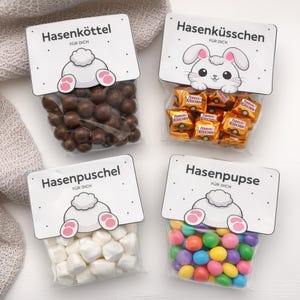 May include: Four clear cellophane bags filled with treats, each labeled with German text and a bunny-themed design. The bags contain chocolate-covered candies, wrapped chocolates, marshmallows, and colorful gumballs. The text on the labels reads "Hasenköttel", "Hasenküsschen", "Hasenpuschel", and "Hasenpupse".