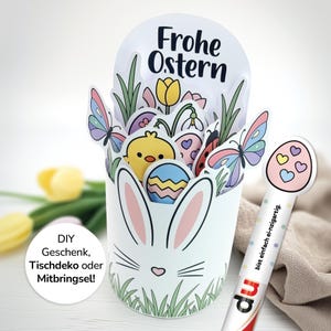 Könnte beinhalten: Ein weißes Oster-Bastelobjekt mit Hasengesicht-Design, gefüllt mit bunten Eiern, Schmetterlingen und einem Küken. Die Worte "Frohe Ostern" stehen oben. Rechts ist ein dekorativer, eiförmiger Stab.