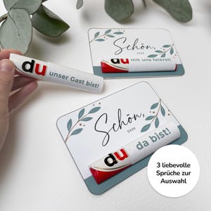 Puede incluir: Barras de chocolate blancas y rojas con la palabra "du" impresa, presentadas con tarjetas decorativas. Las tarjetas tienen la palabra "Schön" y un diseño floral. También se ve el texto "unser Gast bist!".
