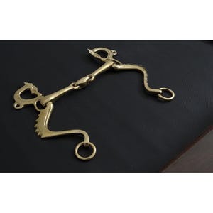 May include: A gold-coloured horse bit with a curved design. The bit has a central joint and rings on each end. The design includes decorative elements and a screw-like detail. The bit is made of metal and is designed for equestrian use.