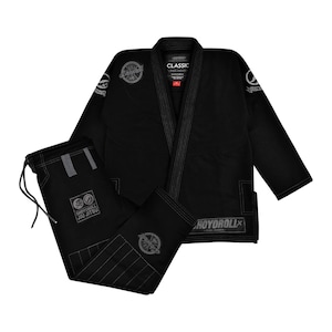 Op de afbeelding: Een zwarte jiu-jitsu gi-set bestaande uit een jas met lange mouwen en een bijpassende broek. De jas is voorzien van een klassieke kraag, Shoyoroll-branding en ronde patches. De broek heeft een taille met trekkoord en een rechthoekige patch.