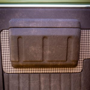 May include: Close-up of a brown car door panel with a textured surface. The panel features a built-in, rectangular storage compartment and a houndstooth pattern accent. The panel is dark brown with vertical lines.
