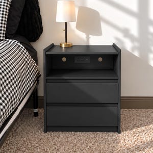 May include: A dark gray nightstand featuring an open top shelf with a built-in power outlet and two lower drawers. A small table lamp with a gold base and white shade sits on top of the nightstand next to a bed with black and white bedding.