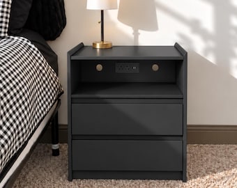 May include: A dark gray nightstand featuring an open top shelf with a built-in power outlet and two lower drawers. A small table lamp with a gold base and white shade sits on top of the nightstand next to a bed with black and white bedding.