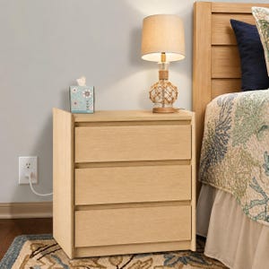 May include: A light wood nightstand with three drawers, topped with a table lamp featuring a tan fabric shade and a rope-wrapped base. A square tissue box with a blue and white floral pattern sits on the surface next to the lamp.