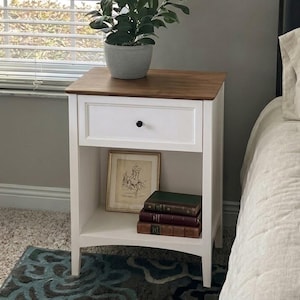May include: A white wooden nightstand with a dark brown wood top, featuring one drawer with a small black knob and an open lower shelf. A potted green plant sits on top, while a framed picture and three stacked books rest on the lower shelf.