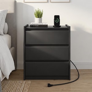May include: A black three-drawer nightstand with a flat top surface. On the nightstand sits a small potted green plant, a book titled The Kinfolk Home, and a black wireless charging station with a visible green charging indicator light.