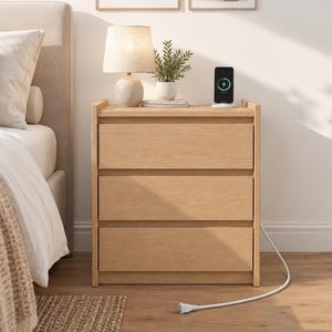May include: A light wood three-drawer nightstand featuring a flat top surface, a white table lamp with a fabric shade, a small potted green plant, and a black smartphone charging wirelessly on a white circular charging pad.