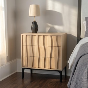 May include: A light wood three-drawer chest featuring vertical wavy textured drawer fronts and a black metal base. A table lamp with a black geometric base and a beige fabric drum shade sits on top of the chest, which is positioned in a room.