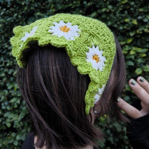 May include: A lime green crochet headband with white and yellow daisy appliqu&eacute;s. The headband has a scalloped edge and is worn on the head. The crochet work is visible and the daisies are evenly spaced.