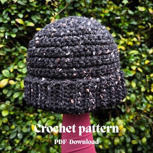 May include: A dark gray crochet hat with flecks of white and brown. The hat has a folded brim and a textured appearance. The text "Crochet pattern PDF-Download" is visible at the bottom of the image.