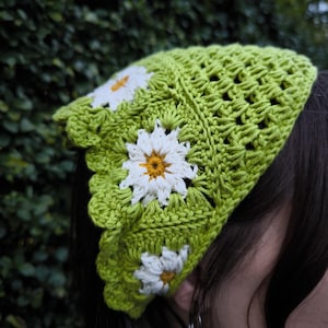 May include: A lime green crochet head covering with three white daisy appliqu&eacute;s with yellow centers. The head covering has a scalloped edge and a textured, open weave design. The background is a blurred green.