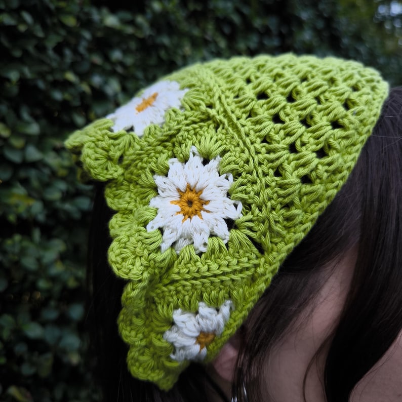 May include: A lime green crochet head covering with three white daisy appliqu&eacute;s with yellow centers. The head covering has a scalloped edge and a textured, open weave design. The background is a blurred green.