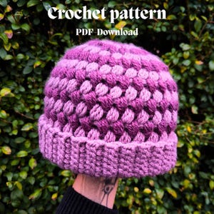 May include: A handmade crochet hat in shades of purple. The hat features a textured, bobble stitch design on the crown and a ribbed brim. The text "Crochet pattern PDF Download" is at the top. The hat is held up against a background of green foliage.