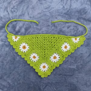 May include: A green crochet bandana with white and yellow daisy flower accents. The bandana has a scalloped edge and tie closures. The handmade accessory is displayed on a gray surface.