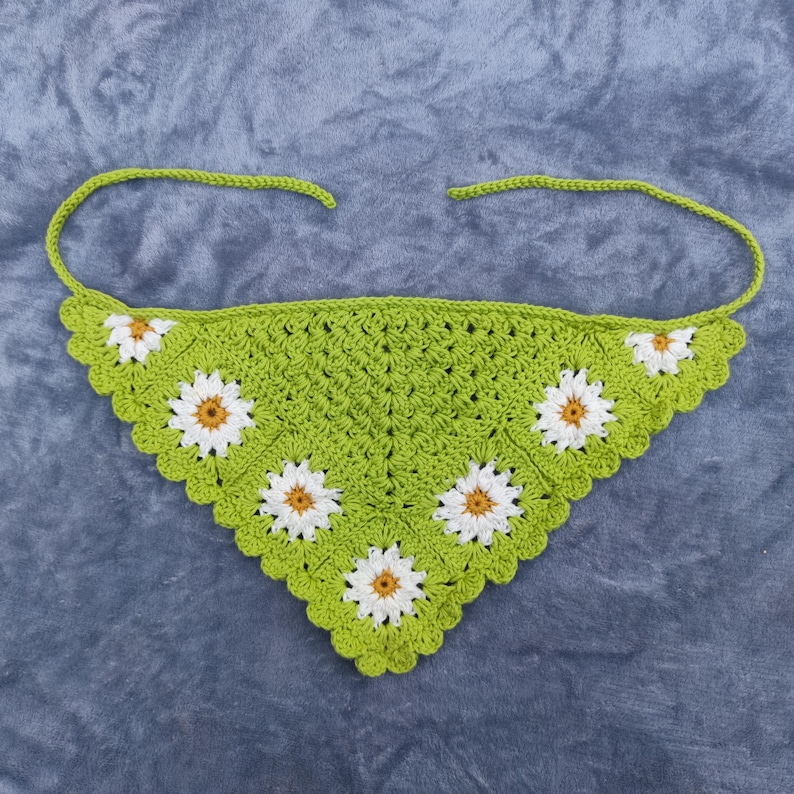 May include: A green crochet bandana with white and yellow daisy flower accents. The bandana has a scalloped edge and tie closures. The handmade accessory is displayed on a gray surface.