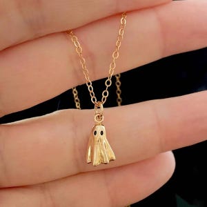 May include: A gold-coloured necklace with a small ghost pendant. The ghost has two black eyes and a draped body. The chain is delicate and also gold-coloured. The necklace is a Halloween-themed accessory.
