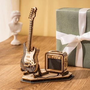 May include: A detailed wooden model of an electric guitar and amplifier. The guitar is light brown and grey, with a white pickguard. The amplifier is a small, square box with a speaker grill. A green gift box with a white ribbon is in the background.