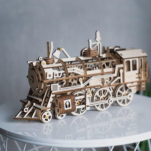May include: A light-coloured wooden model train featuring intricate mechanical gears, a detailed locomotive engine, and spoked wheels. The structure displays a complex assembly of interlocking wooden components, including a cab, boiler, and cowcatcher.