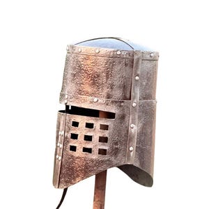May include: An antique metal helmet with a rectangular face guard with square openings. The helmet is riveted and mounted on a rusty metal stand, suggesting historical or medieval themes. The color is a muted, aged brown. The helmet is displayed on a stand.