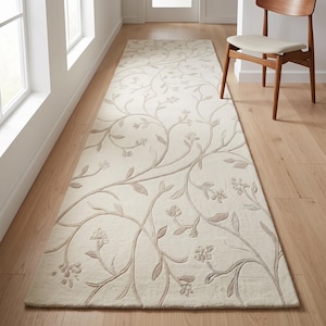 May include: A long, rectangular runner rug in a cream color featuring a raised, tonal vine and floral pattern. The design consists of flowing, curved stems with small leaves and clusters of blossoms that extend across the entire surface.