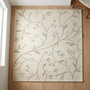 May include: A square area rug in a cream color featuring a raised, tufted vine and floral pattern in a light beige tone. The design consists of sweeping, curved stems with small leaves and clusters of blossoms distributed across the surface.