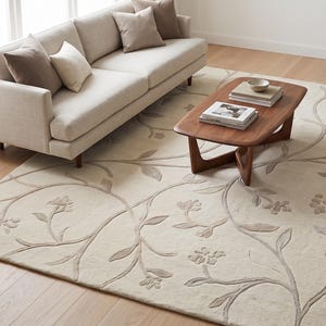 May include: A large rectangular area rug featuring a cream-colored background with a raised, light taupe vine and floral pattern that curves across the surface. The rug has a soft, textured appearance with a consistent botanical design throughout.