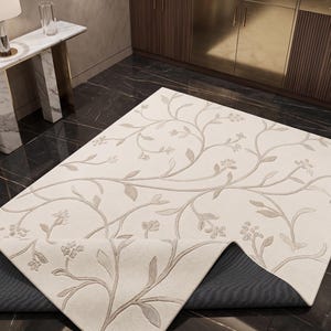 May include: A rectangular area rug featuring an off-white surface with a raised, light beige floral vine pattern. One corner of the rug is folded over to reveal a dark gray, textured backing material underneath the primary light-colored fabric.