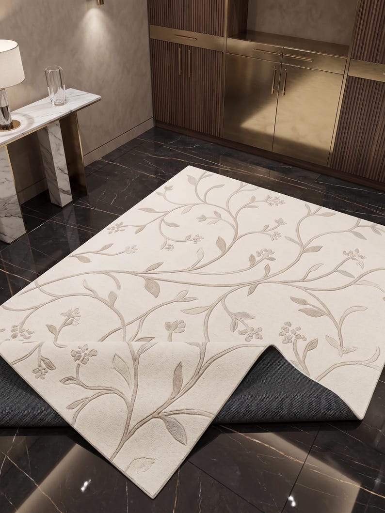 May include: A rectangular area rug featuring an off-white surface with a raised, light beige floral vine pattern. One corner of the rug is folded over to reveal a dark gray, textured backing material underneath the primary light-colored fabric.