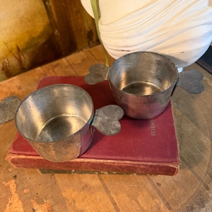 May include: Two small, antique metal bowls with heart-shaped handles. The bowls are sitting on a red book. The bowls have a silver finish and show signs of wear, suggesting age. The background includes a white bust and a painting.