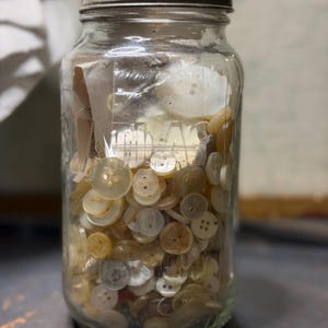 May include: A clear glass jar filled with a variety of vintage buttons in shades of white, cream, and tan. The jar has a metal lid and is sitting on a dark surface. The buttons are of different sizes and shapes.