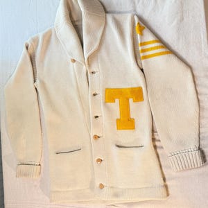 May include: A cream-colored knit cardigan with a shawl collar and button-front closure. The garment features a large yellow felt letter T on the left chest, a yellow star on the left shoulder, and three yellow horizontal stripes on the left sleeve.