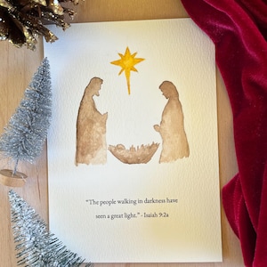 Pode incluir: Cartão de Natal em aquarela com uma cena de natividade com uma estrela, duas figuras e uma manjedoura. O cartão inclui o texto "O povo que andava nas trevas viu uma grande luz." - Isaías 9:2a.