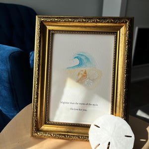 May include: A framed watercolor print with a gold ornate frame. The print depicts a blue wave, seashells, and the text "Mightier than the waves of the sea is His love for you." A white sand dollar rests on the table.