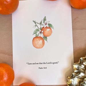 以下が含まれることがあります： 緑の葉と白い花が咲く枝に2つのオレンジが描かれた水彩画プリント。詩篇34:8の「主の恵みを味わい、見よ」というテキストが含まれています。プリントの周りにはいくつかのオレンジがあります。