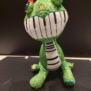 Può includere: Un peluche verde a forma di dinosauro con bocca a tasti di pianoforte, dettagli rossi e gialli e pancia a righe. Il giocattolo ha un design stravagante, con un'espressione amichevole e un tema musicale unico.