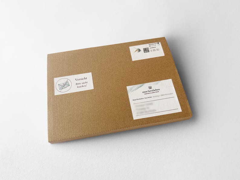 Op de afbeelding: Een bruine rechthoekige kartonnen doos met meerdere witte labels. E&eacute;n label toont een doorgestreepte envelop en de tekst "Vorsicht Bitte nicht knicken!". Andere labels bevatten tekst en een QR-code.