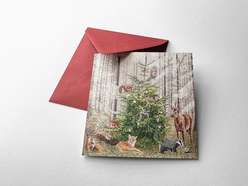 Op de afbeelding: Een kerstkaart met een boslandschap met een versierde kerstboom. Verschillende bosdieren, waaronder vossen, herten en een das, zijn rond de boom afgebeeld. Een rode envelop is zichtbaar op de achtergrond.