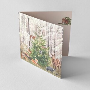 Op de afbeelding: Een gevouwen wenskaart met een bossc&egrave;ne met een versierde kerstboom. De illustratie bevat verschillende bosdieren zoals herten, vossen, eekhoorns en een das. De kaart heeft een lichtbeige binnenkant.